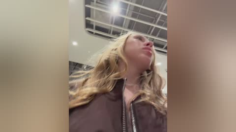 Video of a blonde woman with wavy hair, wearing a dark jacket, standing in a modern indoor space with a high ceiling and exposed beams.