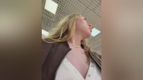 A video of a young woman with long blonde hair, wearing a white blouse and black jacket, looking upward, in a tiled ceiling room.