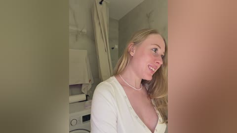 Video of a smiling blonde woman with fair skin and medium-sized breasts, wearing a white cardigan, standing in a beige-walled laundry room.