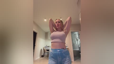 Video of a blonde woman with a curvy physique, wearing a pink crop top and high-waisted jeans, stretching in a modern, minimalist kitchen with light-colored walls and wooden floors.