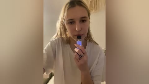 Video of a young Caucasian woman with straight, shoulder-length blonde hair, wearing a white t-shirt, brushing her teeth with an electric toothbrush, in a dimly lit bathroom with beige walls and a tasselled towel hanging.