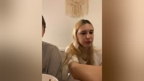 A video captures a blonde woman with fair skin in a white top, looking contemplative, seated next to a man in a grey sweater. The background features a beige macram\u00e9 wall hanging and neutral-colored walls.