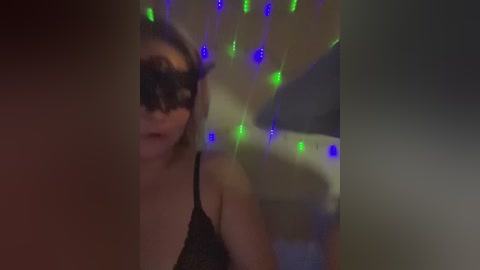 Video of a woman with fair skin, wearing a black lace mask, green and blue fairy lights, and a black bra, blurred background.