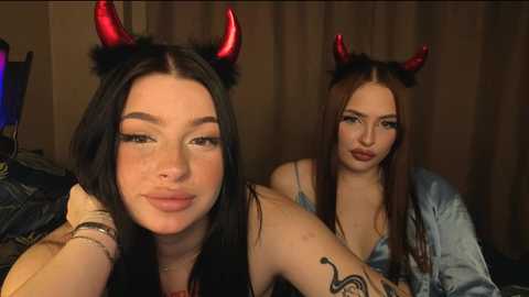Video of two young women with fair skin, long dark hair, and red devil horns. One wears a black bra, the other a light blue top. They're indoors, smiling, with a relaxed, casual atmosphere.
