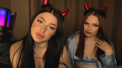 Video of two young women with long dark hair, wearing red devil horns and light blue denim shirts. One has a tattoo on her chest, both have light skin tones, smiling playfully.