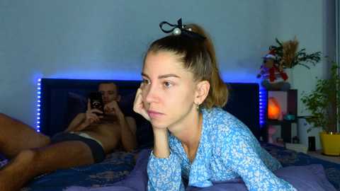 Video of a young woman with fair skin, dark hair tied back, wearing a blue floral shirt, lying on a bed, looking contemplative. A shirtless man lies next to her, partially visible.