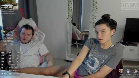 A video of a young man in a white hat, wearing a white t-shirt, sitting in a gaming chair, and a young woman with dark hair, wearing a grey graphic t-shirt, sitting on a sofa, both in a modern living room.