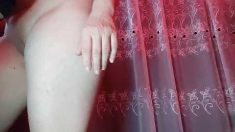 A video of a nude, fair-skinned person with a small breast, standing next to a lace curtain with floral patterns. The individual's hand rests on their hip. The setting appears to be indoors, with soft lighting.