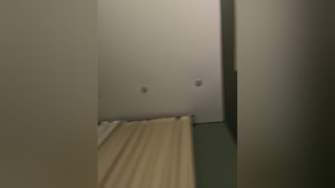 Video of a narrow, dimly lit hallway with a beige wall, a light wooden floor, and a closed door on the right. The floor is slightly uneven, and the lighting is soft, creating a somber atmosphere.