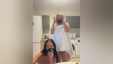 Video of two women in a modern kitchen. One, with long black hair and wearing a white towel, takes a selfie. The other, with blonde hair, wears a mask and towel.