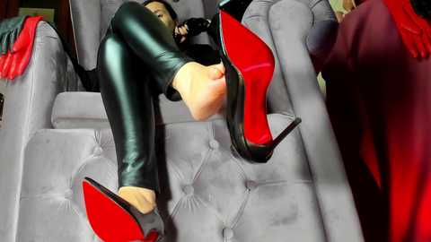 Video of a woman lying on a gray, tufted armchair, wearing shiny black leather leggings and red-bottomed high heels, with a red dress draped over her legs.