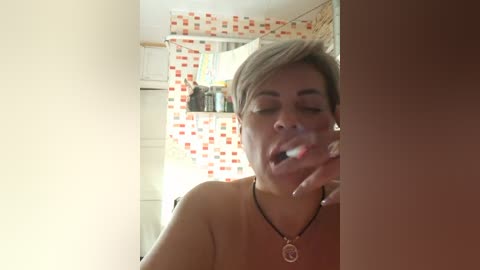 Video of a woman with short, light brown hair and a tan complexion brushing her teeth in a bathroom. She wears a sleeveless orange top and a black necklace with a circular pendant. The background features a tiled shower curtain with red and white squares.