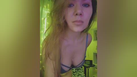 Video of a young woman with long, messy blonde hair, wearing a blue bra, in a dimly lit room with yellow-green tint. She has a neutral expression and is looking directly at the camera.
