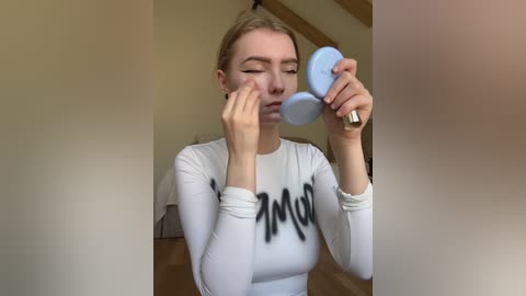 Video of a young woman with fair skin and blonde hair, wearing a white long-sleeve shirt with black text, applying makeup with a blue brush. Background shows a beige wall and wooden floor.