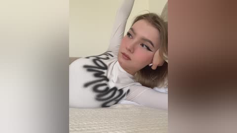 Video of a young Caucasian woman with light skin, blonde hair, and makeup, lying on a beige bed, wearing a white long-sleeve top with black text, looking relaxed and thoughtful.