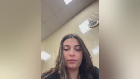 Video of a young woman with long brown hair, wearing a black shirt, standing in a beige-tiled bathroom with fluorescent lights overhead.