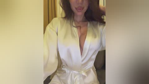 Video of a woman with fair skin and long brown hair, wearing a white satin robe, smiling. Background features a beige curtain and a wooden door.