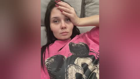 A video of a young woman with fair skin, long dark hair, and red-painted nails, wearing a pink t-shirt with a skull graphic. She's lying on a beige couch, holding her forehead with a distressed expression.