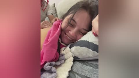 Video of a smiling woman with long hair, wearing a pink blanket, lying on a gray and white striped bed, with a blurred background.