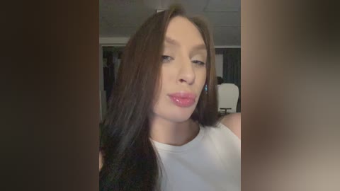 A video of a young Caucasian woman with long, straight brown hair, fair skin, and a white tank top, making a pouty face, set against a blurred, dimly lit room with white ceiling tiles.