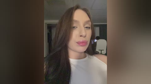 Video of a young woman with long, dark brown hair, fair skin, and full lips. She wears a white sleeveless top. Background features a dimly lit room with a chair and a door.