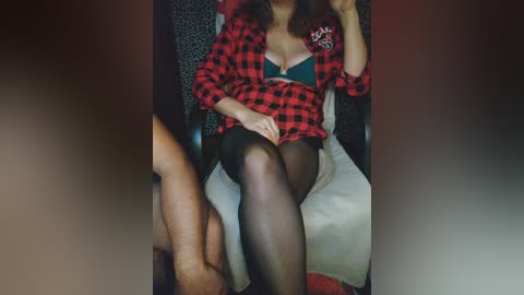 Video of a woman with light skin, wearing a red plaid shirt and black bra, sitting on a chair. Her legs, clad in sheer black stockings, are visible.