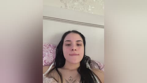 Video of a young woman with long black hair, fair skin, and a small tattoo on her chest, wearing a white lace bra, lying on a bed with pink floral pillows.