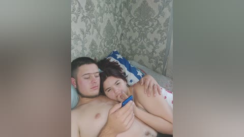 A candid video of a young couple, both topless, lying in bed. The man, with short dark hair, is holding a blue phone, and the woman, with brown hair, smiles. The room has patterned wallpaper and a decorative pillow.