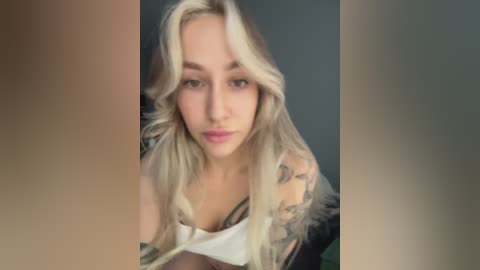 A video of a fair-skinned, blonde-haired woman with wavy hair, wearing a white tank top, revealing a tattoo on her right shoulder. She has a neutral expression and stands against a blurred background.