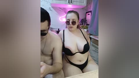 A video of a plus-sized woman with light skin and brown hair, wearing black lingerie, sitting on a bed in a dimly lit room with neon lights and a pink background.