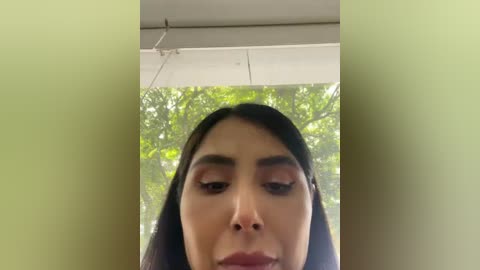 Video of a woman with long dark hair, wearing makeup, looking down. Green filter surrounds her face, creating a soft, dreamy effect. Background shows blurred greenery and a white ceiling.