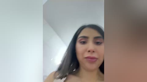 Video of a young Latina woman with medium skin tone, dark hair, and light pink lipstick. She wears bold pink eyeshadow and is smiling. The background features a white wall and a shadowed corner.