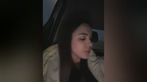 A dimly lit video of a young woman with long, straight black hair, wearing a beige jacket, sitting in a car, eyes closed, appearing relaxed.