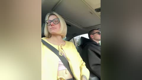 Video of a blonde woman with glasses wearing a yellow jacket in a car, looking relaxed; a man in the background appears to be driving.