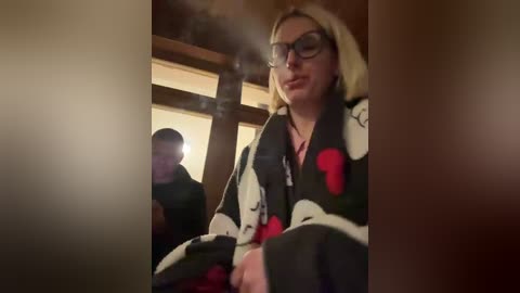 Video of a blonde woman in glasses, wearing a black robe with red and white patterns, sitting on a wooden bench, with a man in the background. The scene is dimly lit, creating a warm, cozy atmosphere.