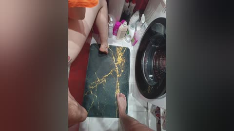 A video showing a woman's bare feet on a black and gold marble mat, standing in a laundry room with white tiles, pink and purple cleaning products, and a black washing machine in the background.