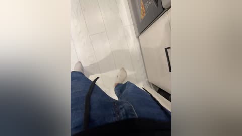 A video showing a person's legs in blue jeans, lying on a white bedsheet in a cramped, dimly lit room. The background reveals a white door and a small, beige cabinet.