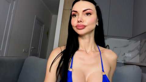 Video of a slim, pale-skinned woman with long black hair, wearing a bright blue bikini top, posing indoors with gray kitchen cabinets and a gray couch in the background.