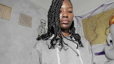 Video of a young Black girl with long, braided hair, wearing a grey hoodie, standing in a cluttered room with abstract art and a skull on the wall.