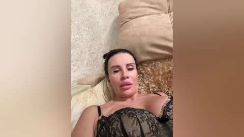 Video of a fair-skinned woman with dark hair, wearing black lace lingerie, reclining on a beige couch with floral cushions. She has a pensive expression with parted lips, and the background includes a textured beige wall.