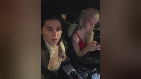 Video of two young women in a dimly lit car, one with dark hair, one with blonde hair, both focused on their phones, wearing casual clothes.