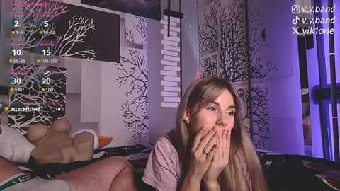 A video captures a blonde woman in a light pink top, covering her mouth in surprise, in a dimly lit room with black-and-white tree art and a bed with a dark blanket.