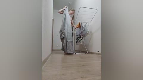 Video of a blonde woman in a white shirt and grey pants, hanging laundry in a narrow hallway with light wooden floors and white walls.