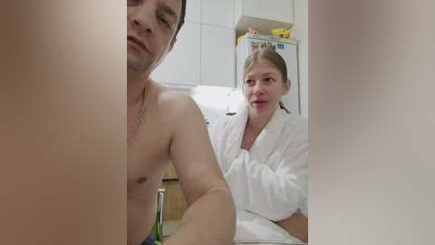 Video of a shirtless man and a woman in a white bathrobe, standing in a kitchen with white cabinets and a yellow refrigerator.