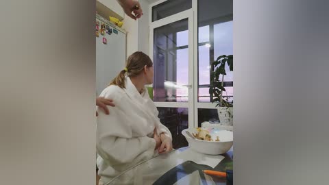 Video of a woman in a white sweater sitting at a kitchen table with a potted plant and a bowl of food. Background shows a refrigerator, window, and a man's hand reaching in.