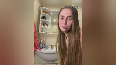 Video of a fair-skinned, nude woman with long, straight brown hair, puckering her lips in a bathroom with beige tiles, white sink, and shelves holding toiletries.
