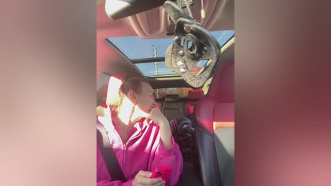 A video of a woman in a pink jacket, seated in a car, with a large, decorative steering wheel hanging from the ceiling, and sunlight streaming through the open roof.