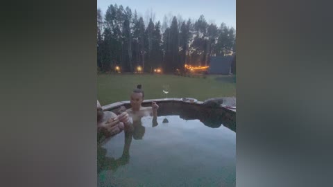 Video of a naked woman with short blonde hair floating in a hot tub in a dark, wooded backyard at twilight.
