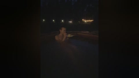 A dimly lit video of a naked man, with a muscular build, sitting in a hot tub at night. He has short, light brown hair and is holding a book. The background features dark, blurry trees and a faintly visible house with lit windows.
