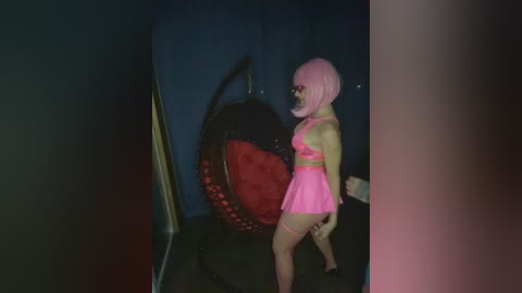 Video of a pale-skinned, petite woman with pink bobbed hair, wearing a pink mini dress, standing in a dimly lit room with a black wicker chair and a red cushion.